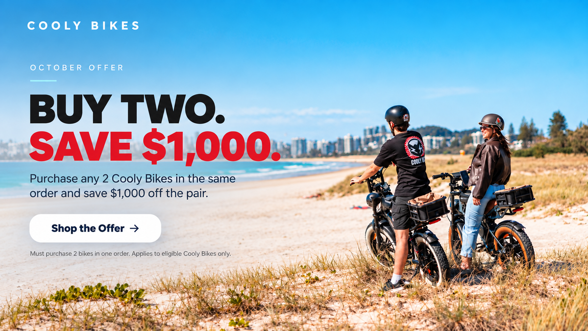 Promotional beach scene for Cooly Bikes showing two riders on electric bikes beside a sandy shoreline under a blue sky, with large text reading 'BUY TWO. SAVE $1,000.